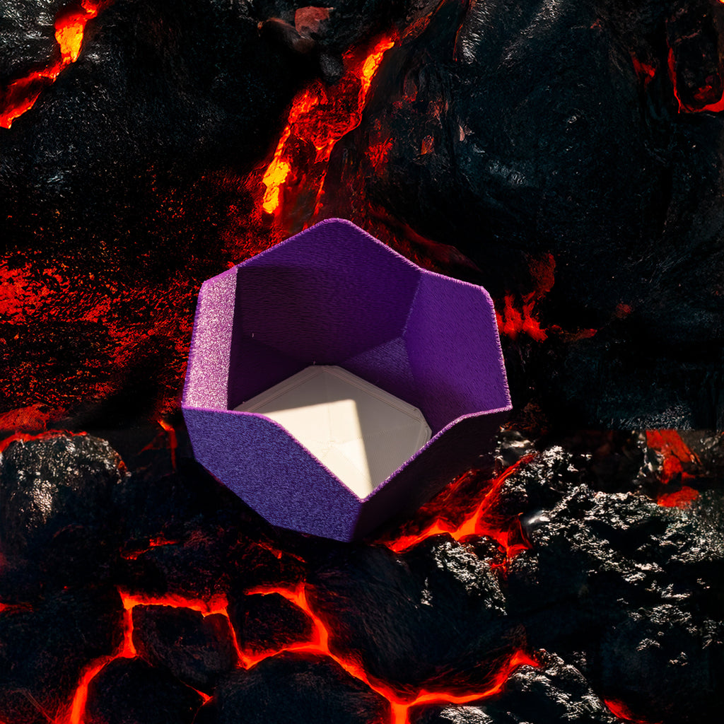 A decorative, geometric, violet catch all with an interchangeable coaster. The coaster inside is in white. The catchall is against rocks cooling from streams of lava.