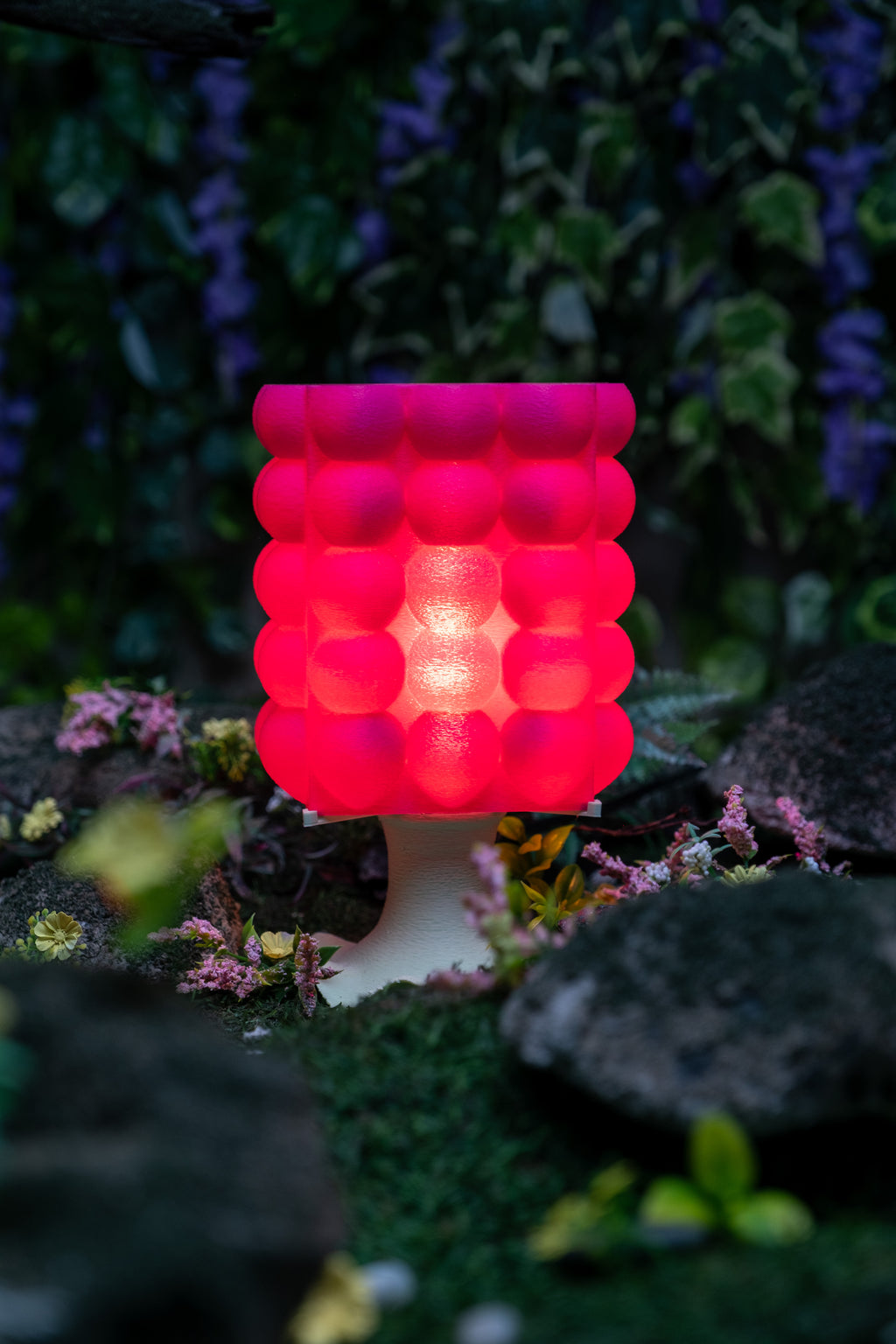 A pink, bubble-textured 3D-printed lamp alight on a lush forest floor.
