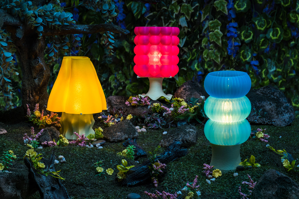 Group photo of the Making Things 3D-printed Lamp Collection. From left to right, there is a yellow, hot pick, and blue lamp placed on top of rocks along the forest floor.