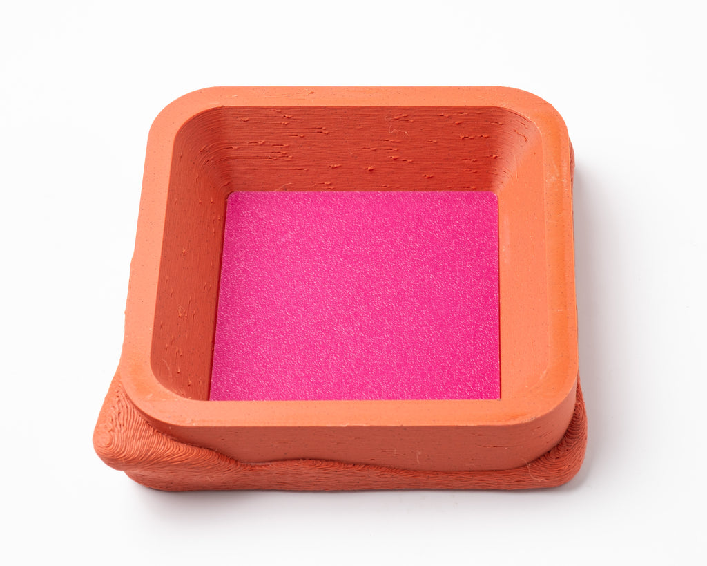 A decorative, square, orange catch all with an interchangeable coaster. The coaster inside is in hot pink. 