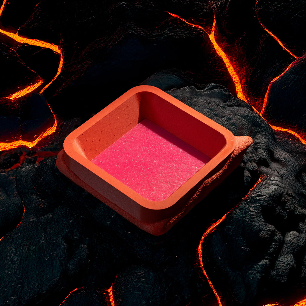 A decorative, square, orange catch all with an interchangeable coaster. The coaster inside is in hot pink. The catchall is against rocks cooling from streams of lava.